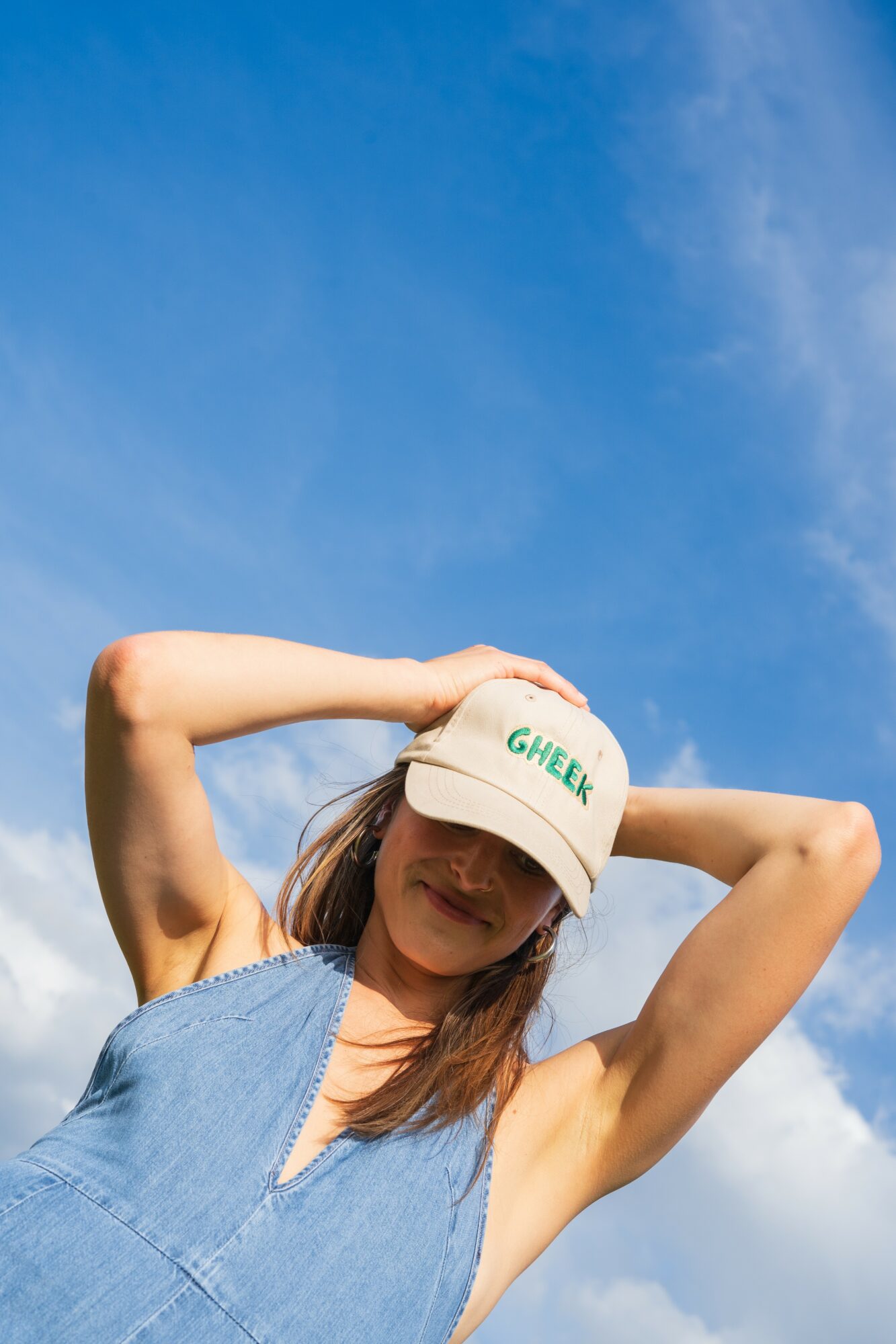 Woman wearing a white cap and blue sleeveless top, standing outdoors against a blue sky with clouds.