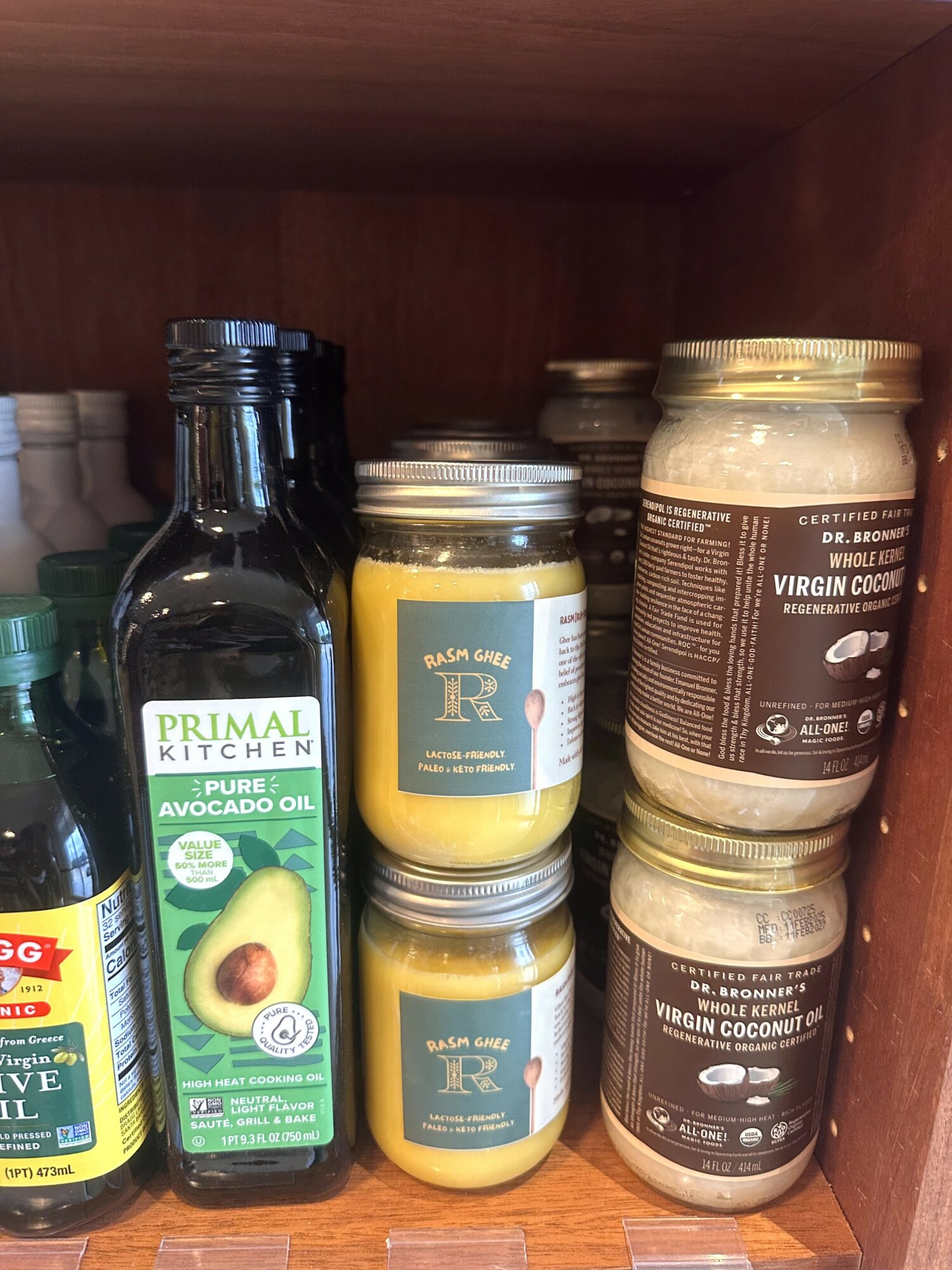 Shelf with jars of coconut oil and containers, and a bottle of avocado oil, all on a wooden shelf.