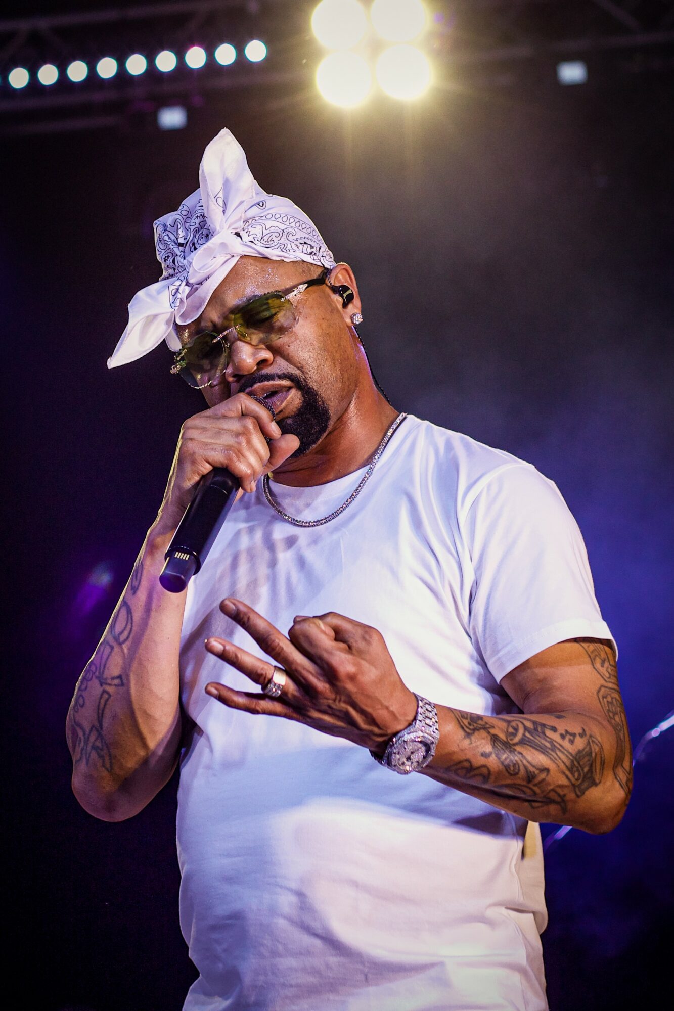 Male performer with bandana, sunglasses, white t-shirt, holding microphone, performing on stage with bright lights behind.