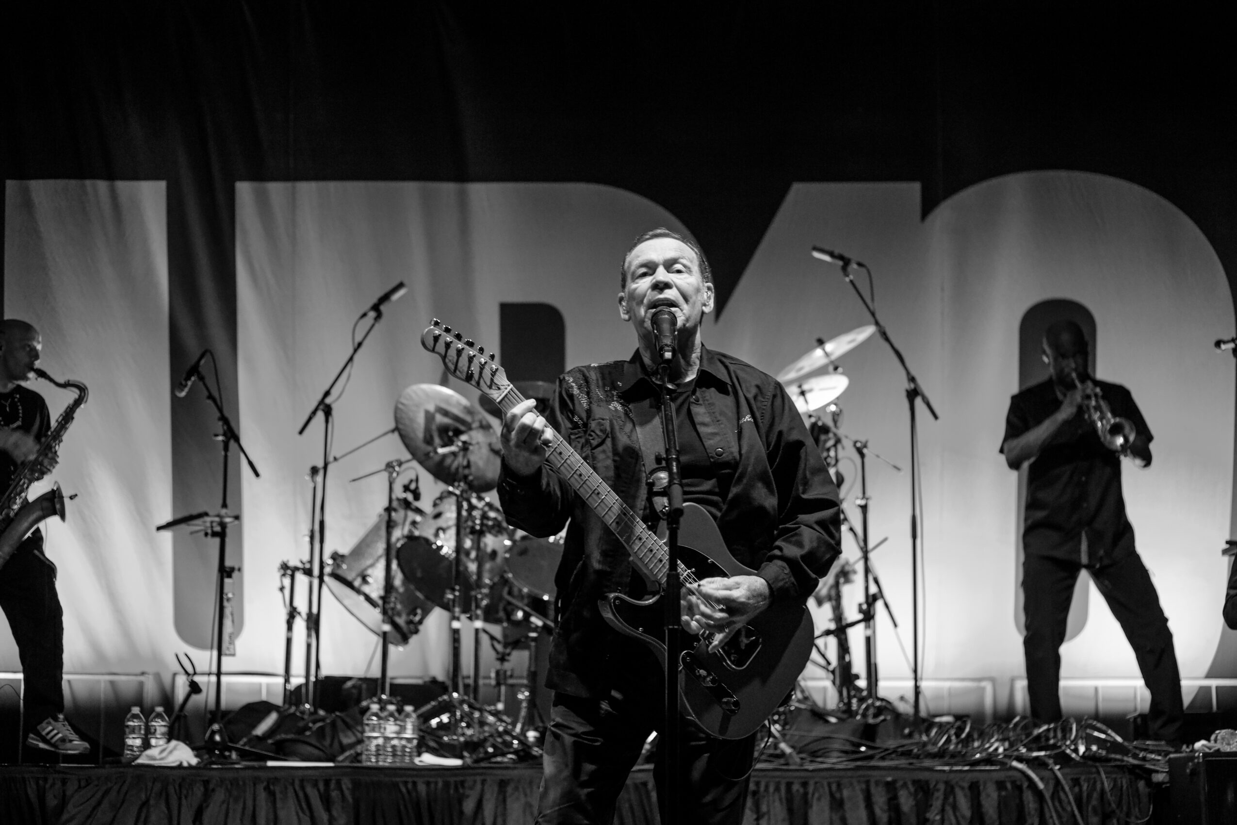 Musician playing guitar on stage with band members, microphones, and large backdrop, in black and white.
