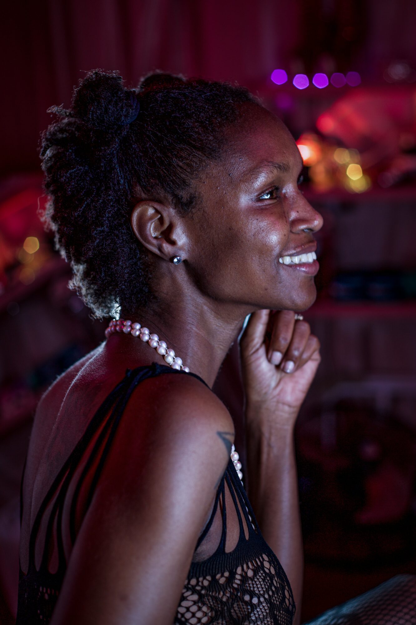 Smiling woman with curly hair wearing a pearl necklace and black dress, in a dimly lit setting.