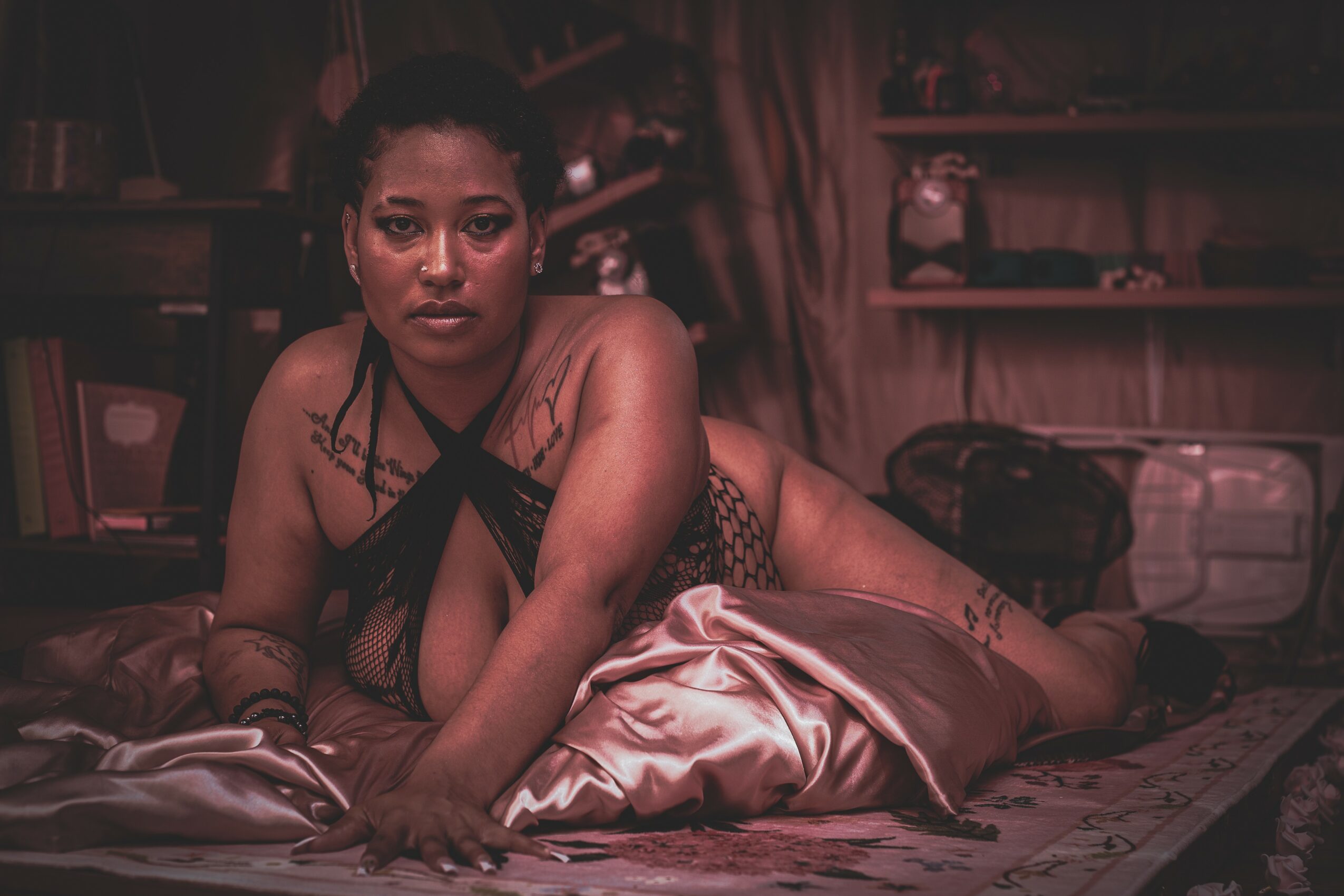 Woman lying on bed with pillows, looking at camera, in a dimly lit room with shelves in background.