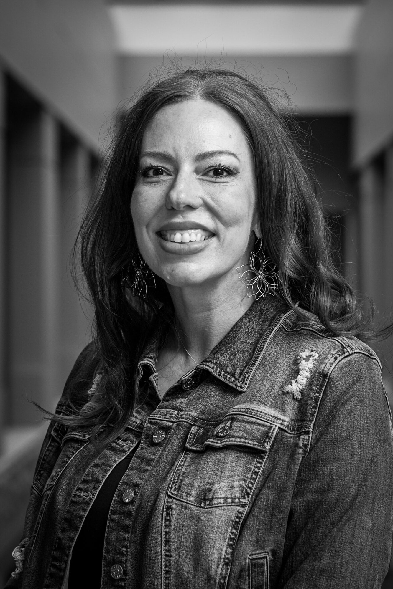 Woman with long hair smiling, wearing a denim jacket, in a hallway setting.