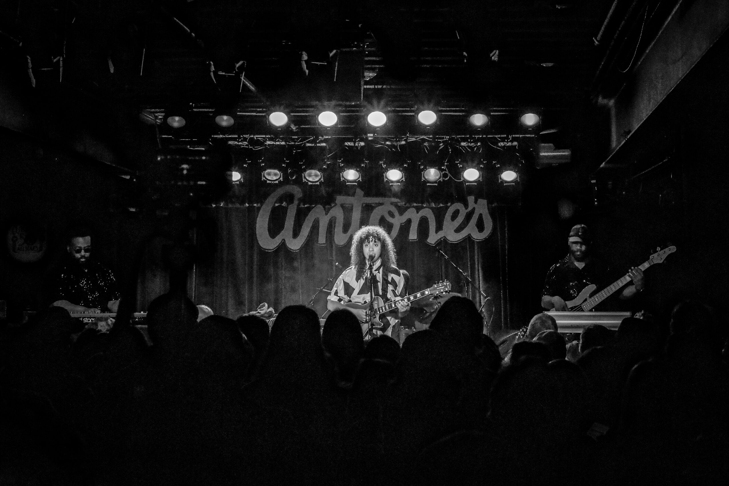 Stage with a performer playing guitar, band members, and audience silhouettes, with a sign reading 'Amores' in the background.