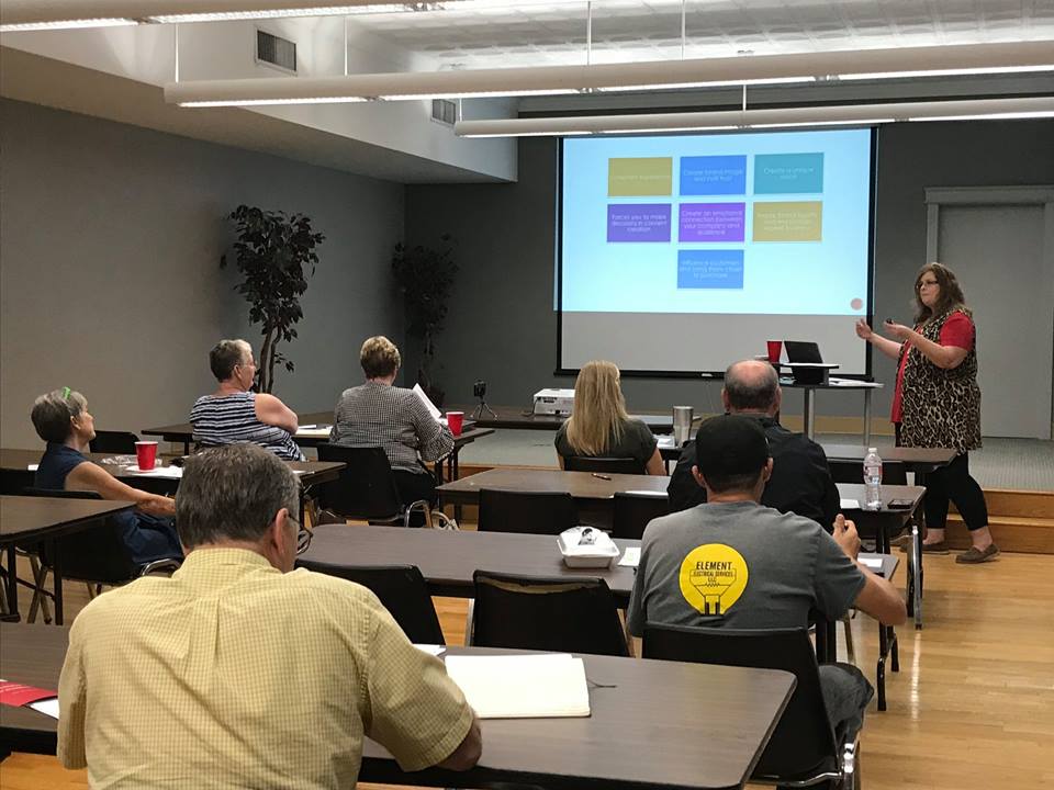 A woman presents to a group in a conference room with a large screen displaying a colorful grid, attendees seated at tables.