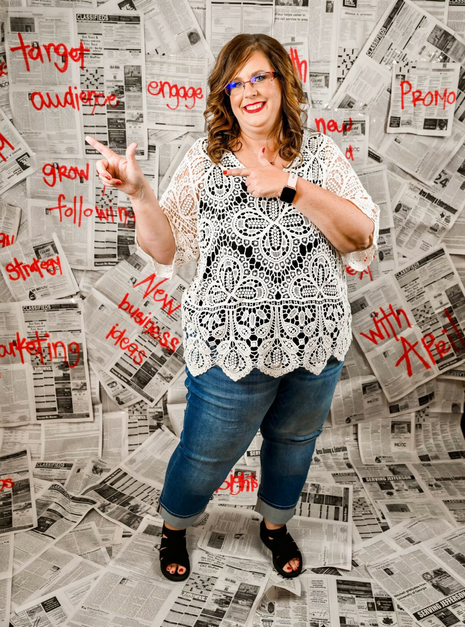 Woman with glasses and curly hair smiling, pointing, standing in front of newspaper background with red handwritten notes.