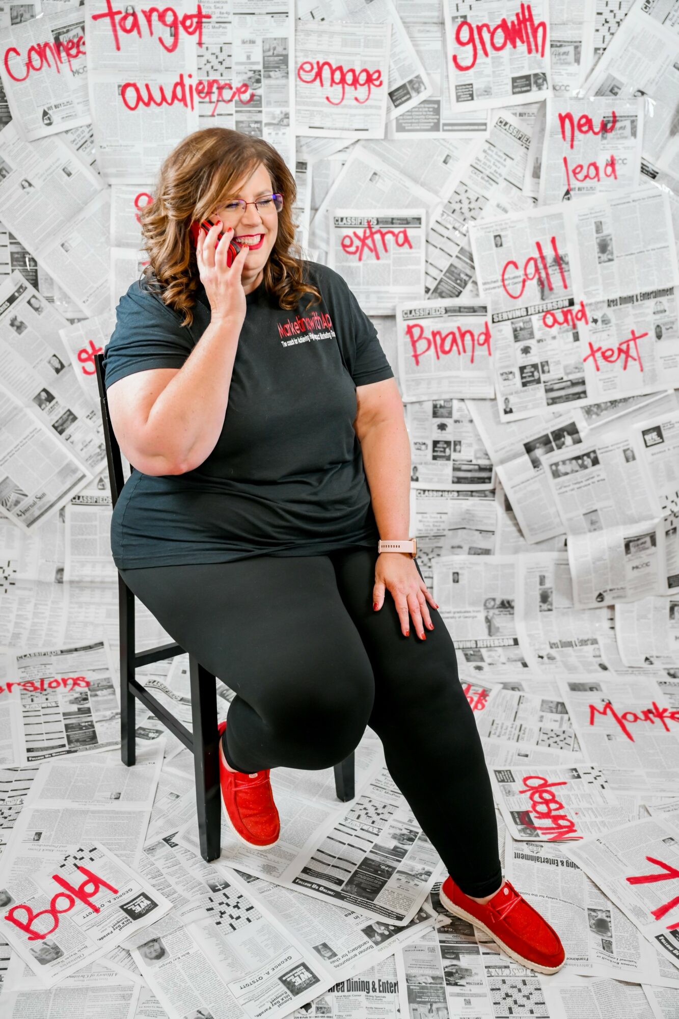 Woman sitting on a stool, smiling, with a wall covered in newspapers and red handwritten words behind her.
