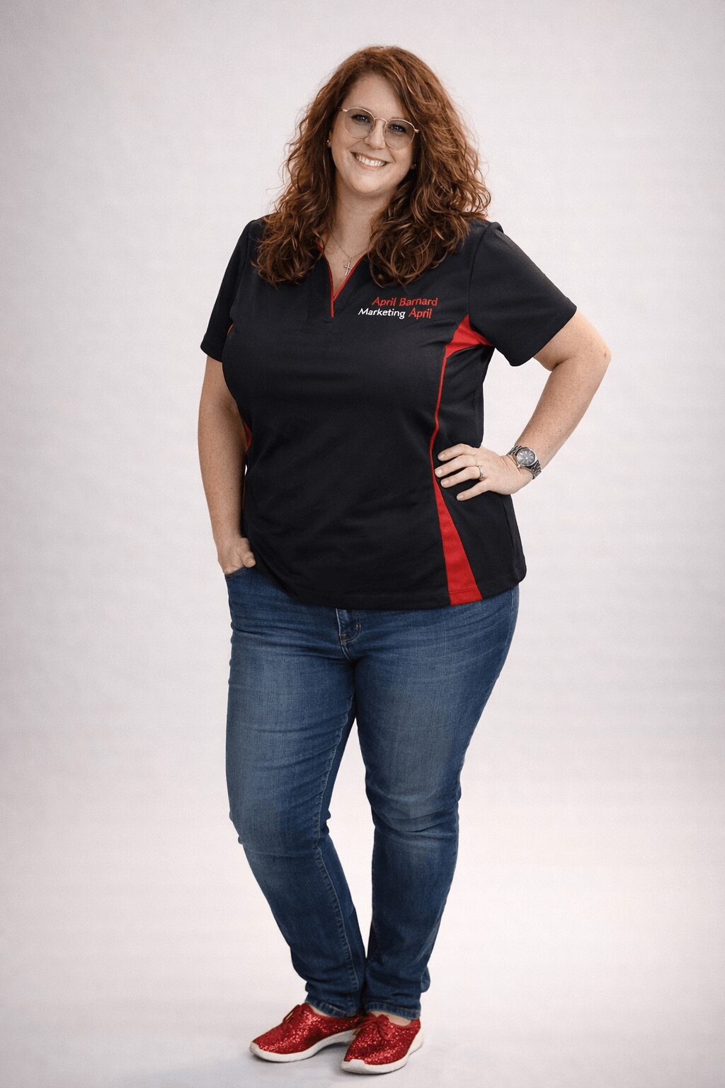 Woman with curly red hair wearing a black and red uniform top and jeans, smiling, standing with one hand on her hip.