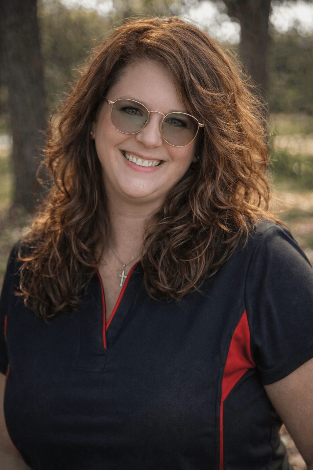 Woman with curly hair wearing glasses and a dark top outdoors, smiling.