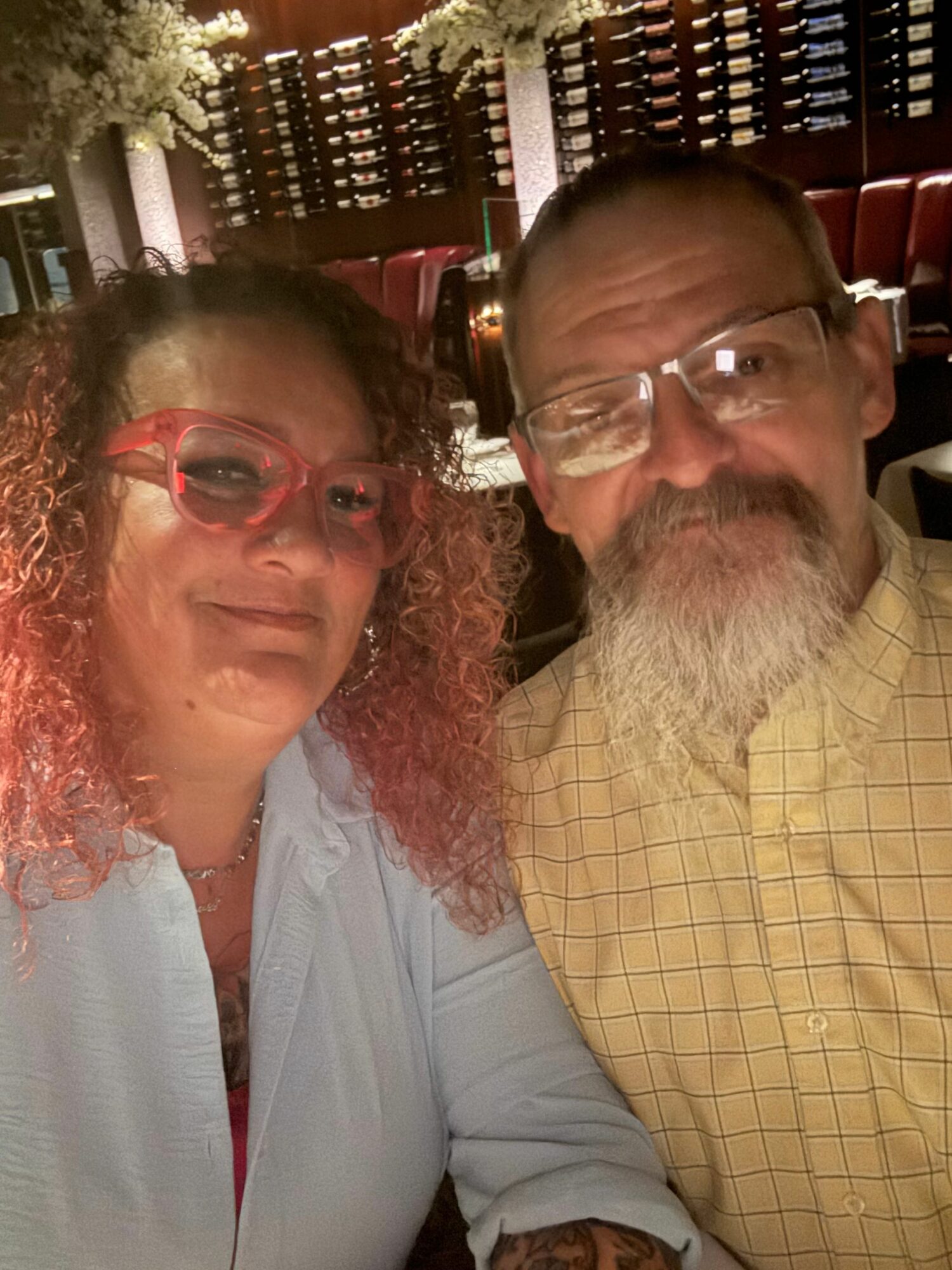 Two people, a man with glasses and a woman with red glasses, pose closely together indoors.