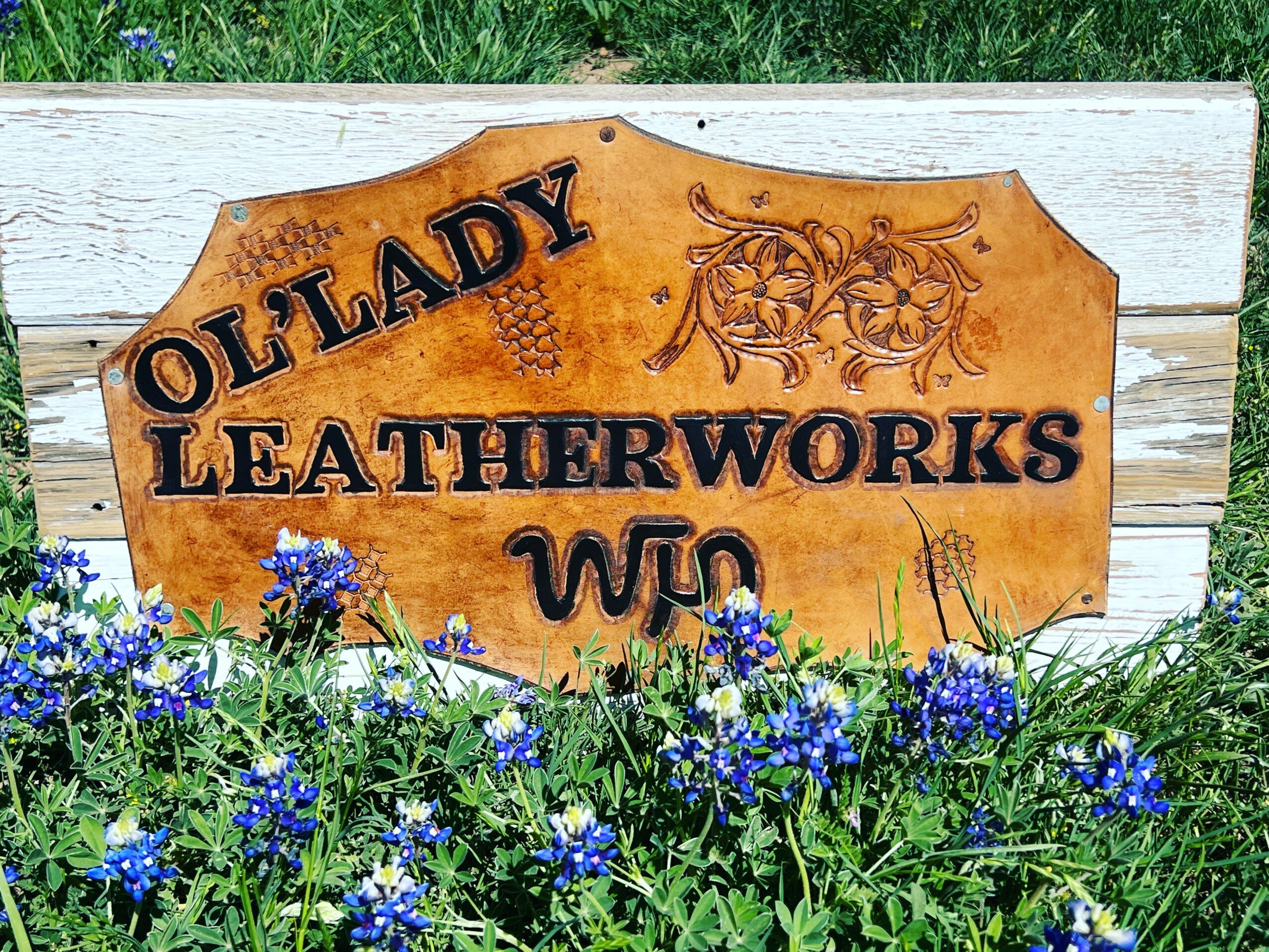 Wooden sign reading 'Ollady Leatherworks' with decorative design, surrounded by blue flowers and green grass.