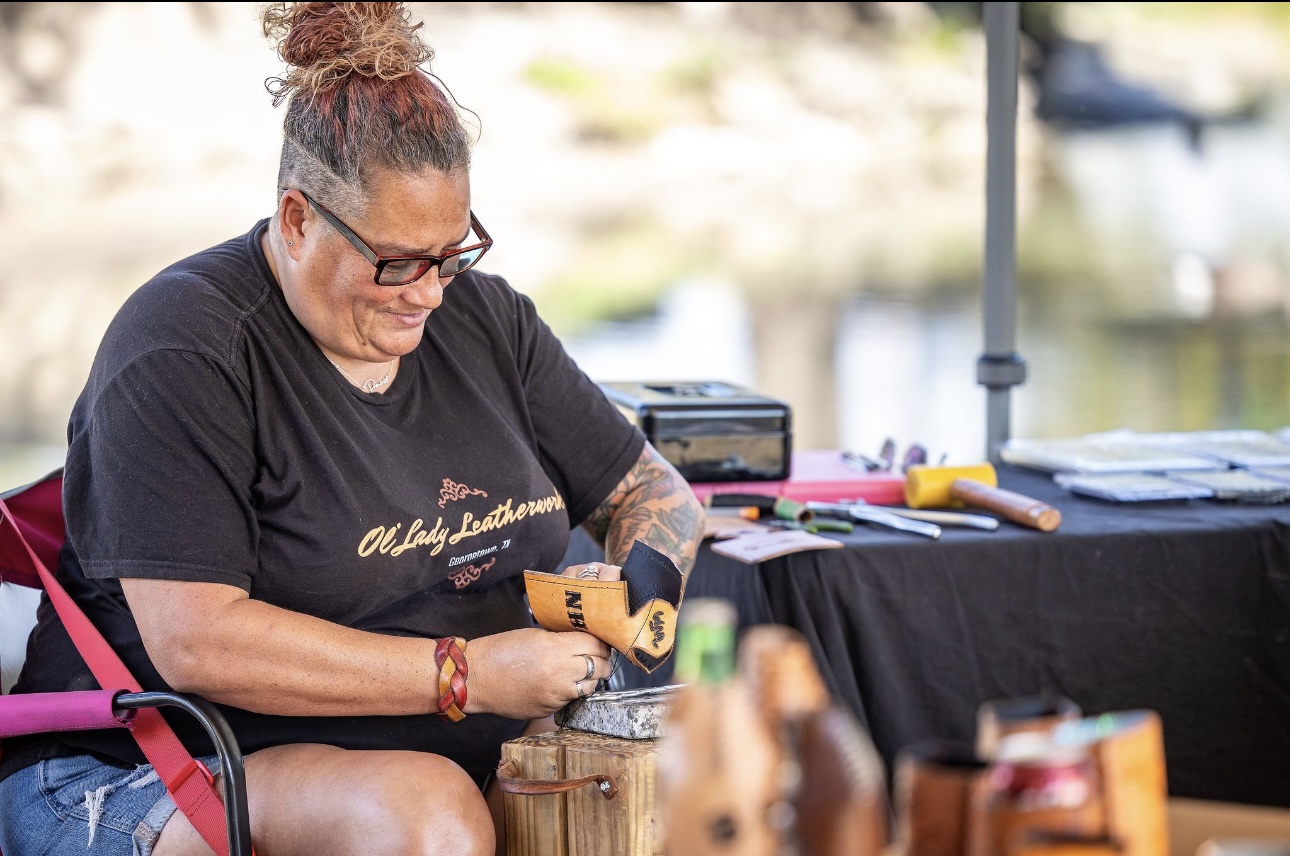 Woman with glasses and curly hair, sitting outdoors, opening a small box, with a table and water in background.