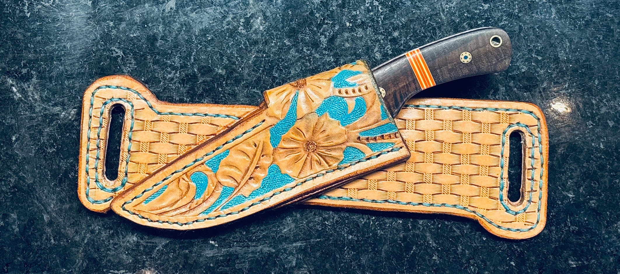 Knife with a floral design handle in a leather sheath on a dark surface.