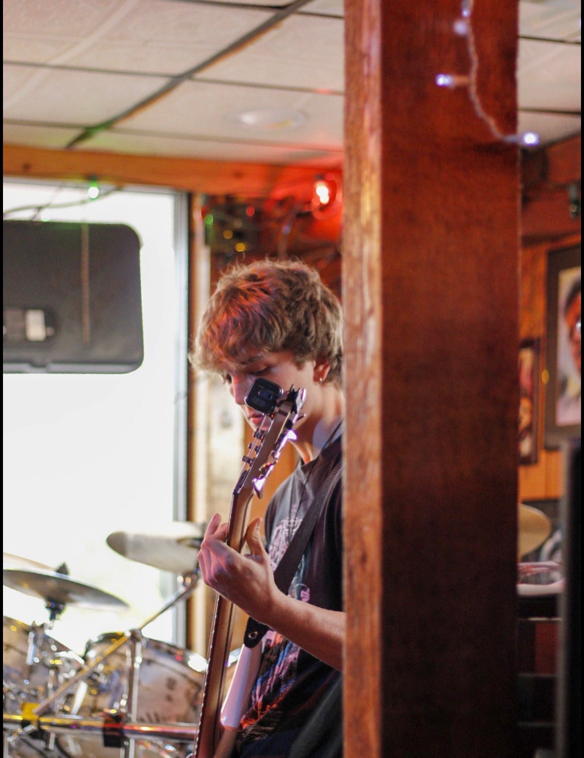 Person with curly hair playing guitar in a room with a drum set and warm lighting, partially obscured by a wooden pillar.