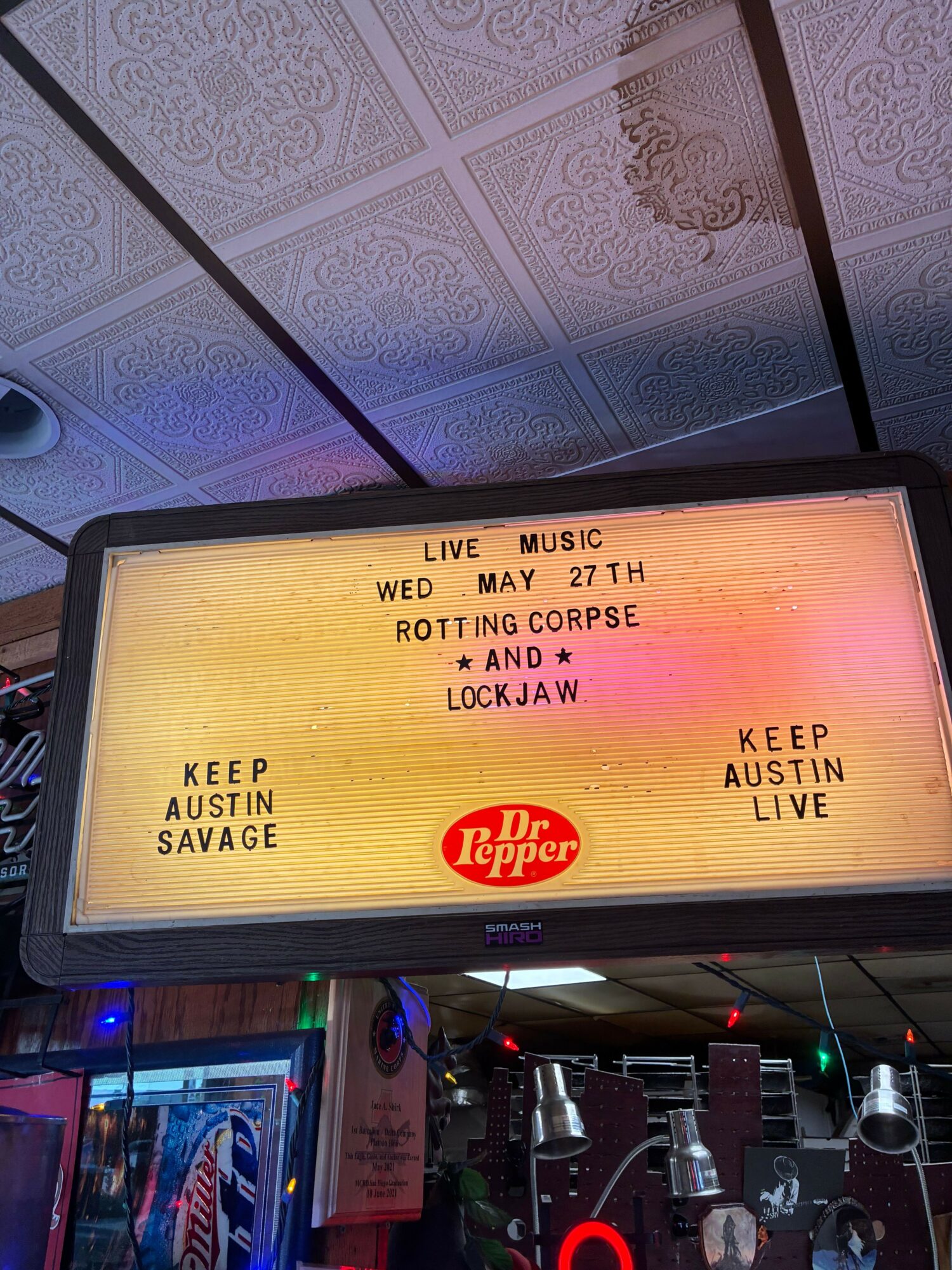 Electronic sign displays event details and a logo, with a bar scene in the background, including bottles and a neon sign.