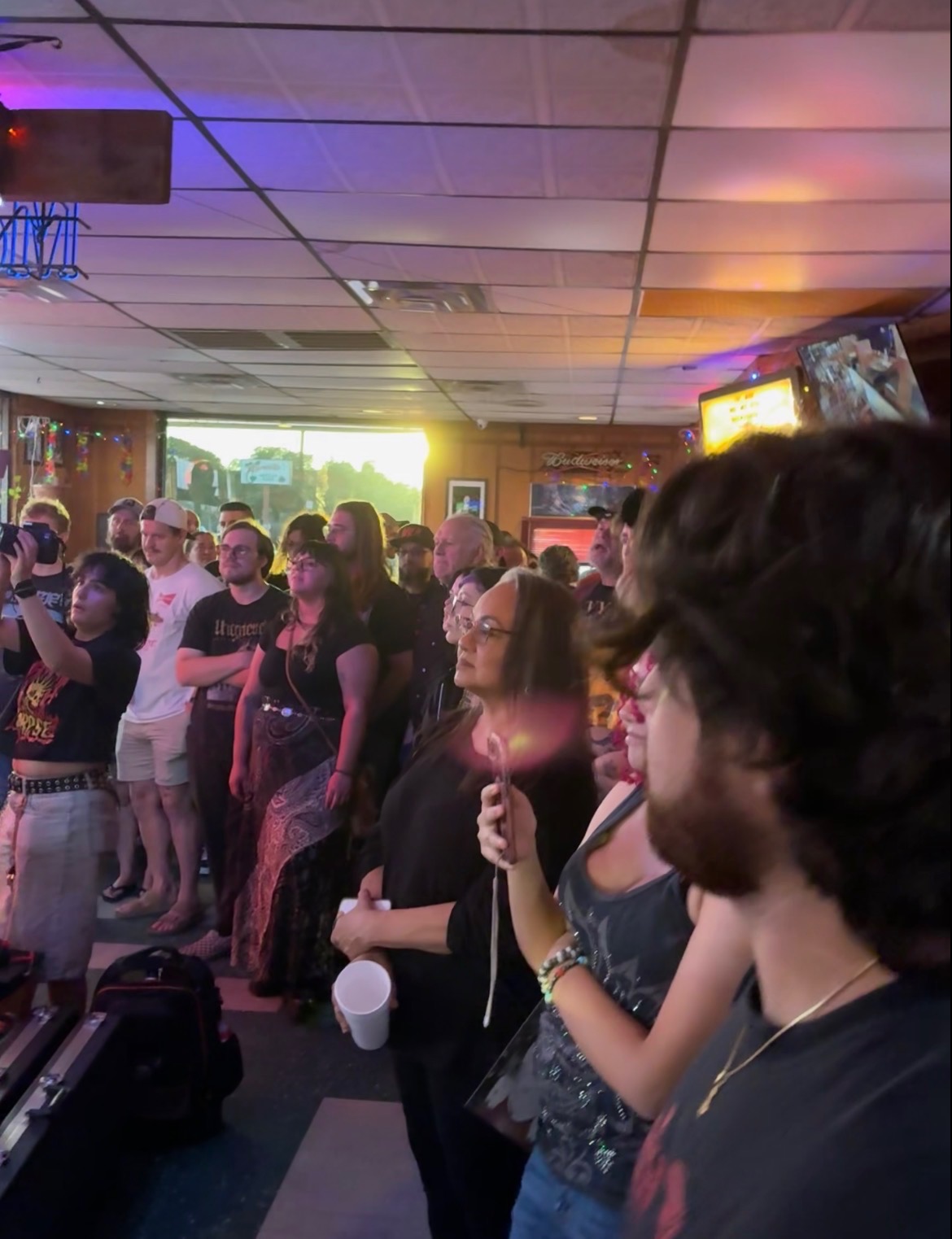 Crowd of people gathered indoors, some holding drinks, with a bright window and television screen in the background.