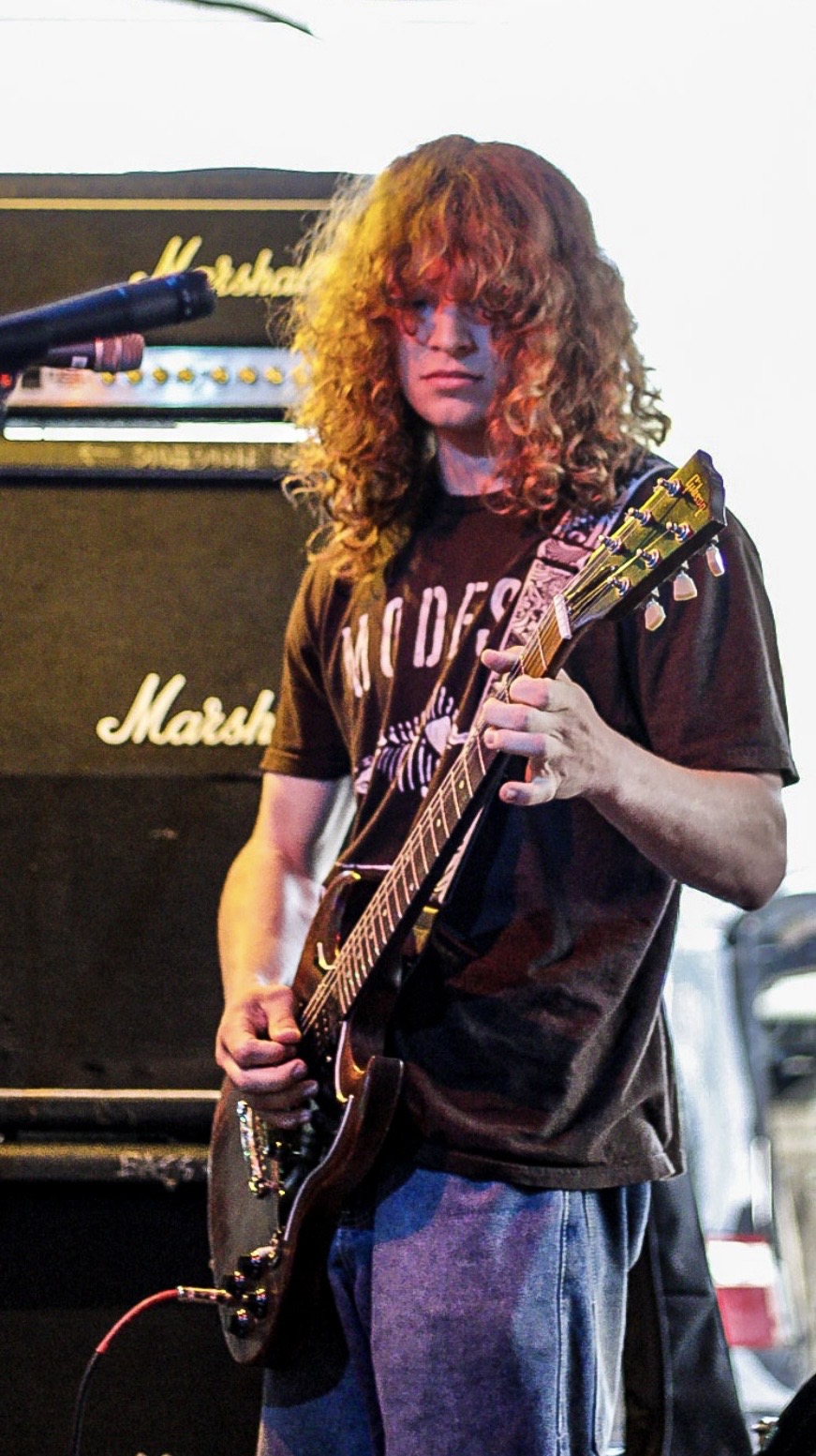 Person with curly hair playing an electric guitar on stage, Marshall amplifier in background.