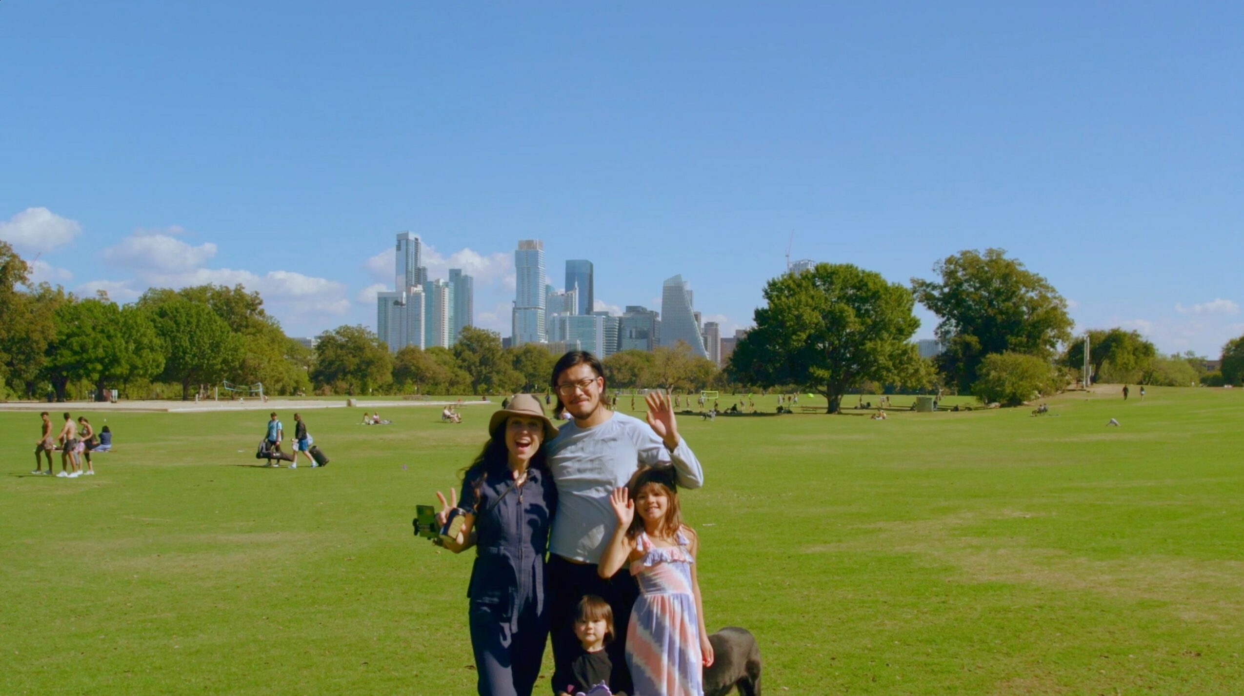 Group of four people standing on a grassy park with city skyline in background, trees, and other park visitors.