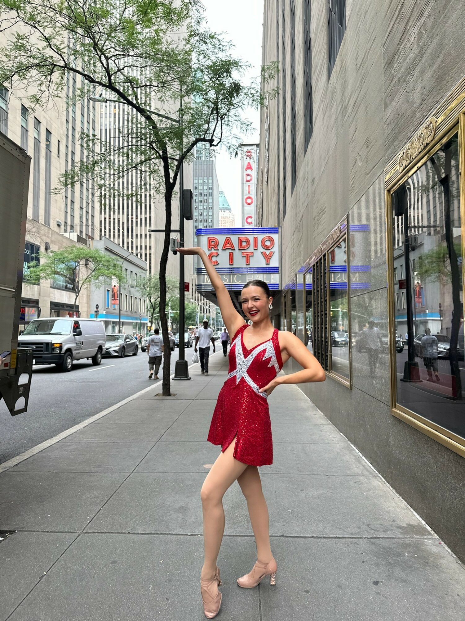 Person in red dress posing on city sidewalk with buildings and trees, radio sign in background.