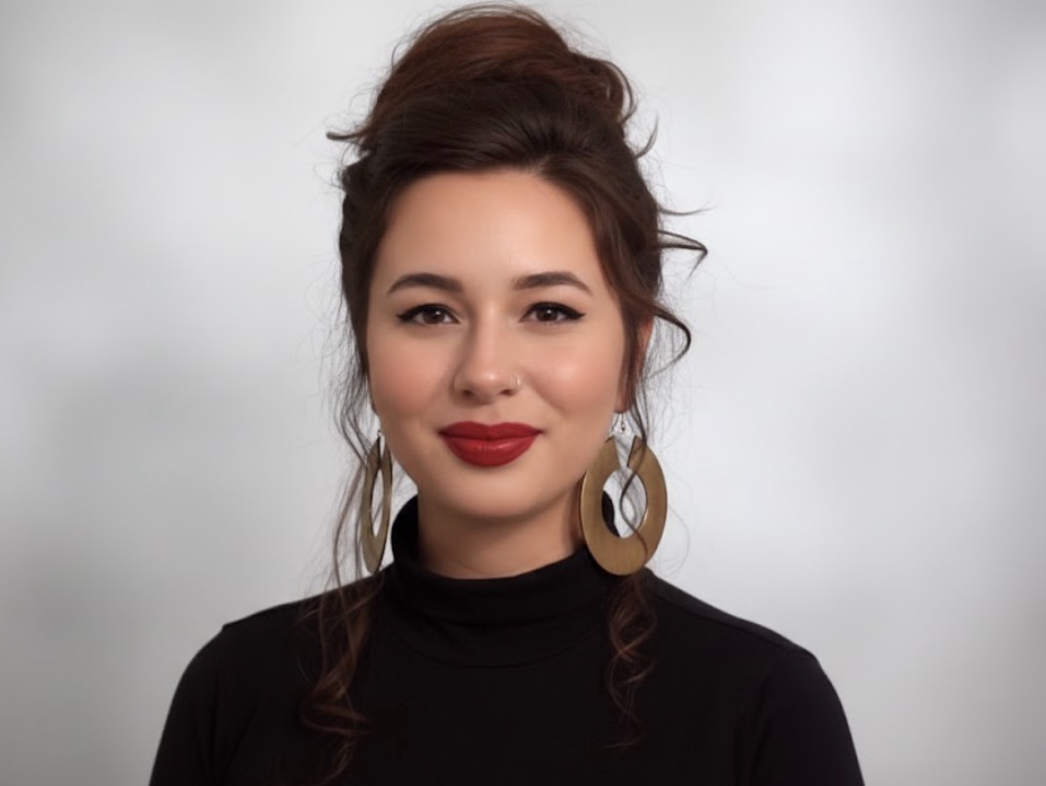 Portrait of a woman with dark hair styled in an updo, wearing large earrings and a black top, smiling.