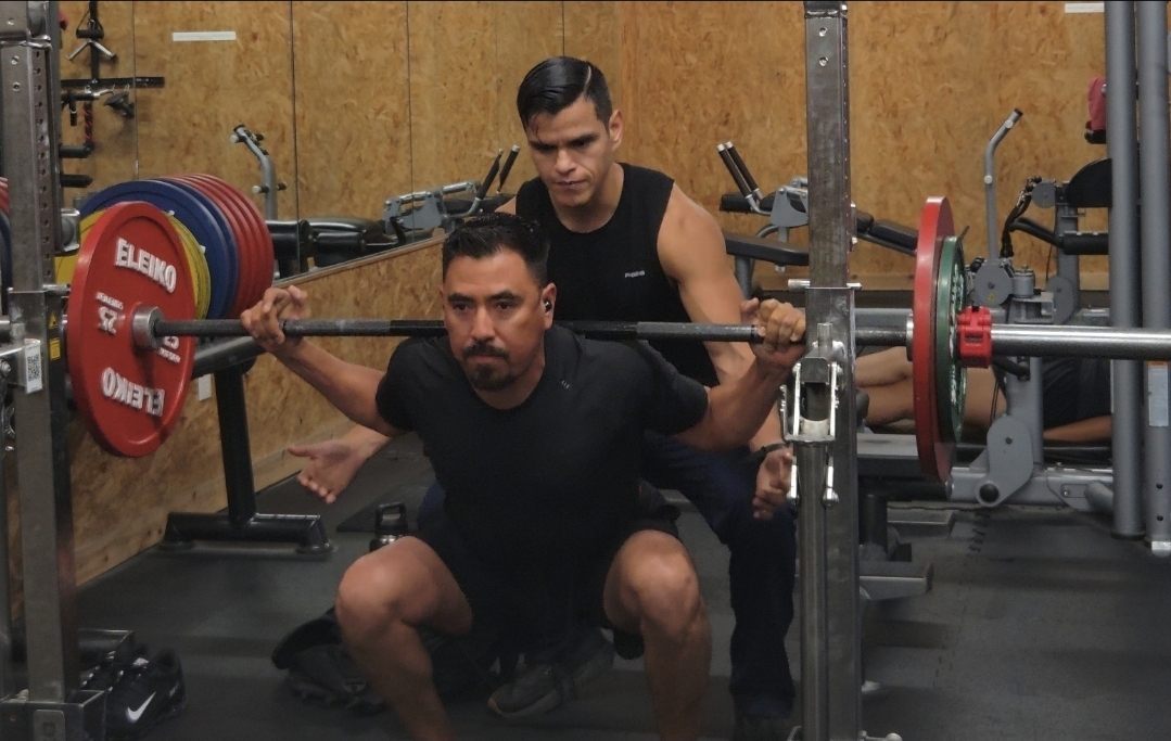 Two men in a gym, one squatting with a barbell, the other standing behind him, spotting him during a workout.