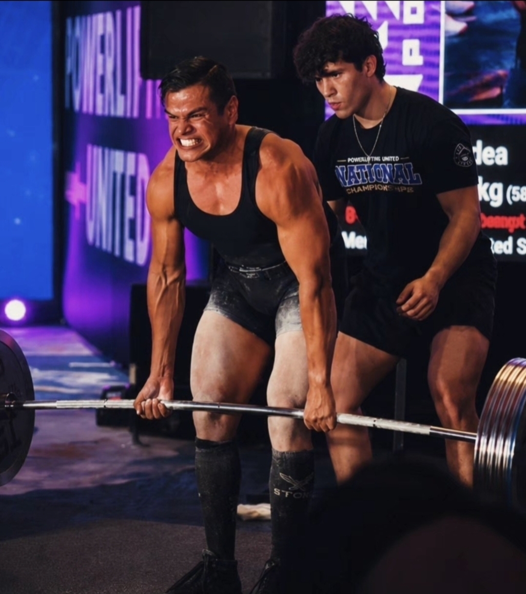 Two men lifting a barbell on stage, one smiling, with a purple-lit background and digital screens.