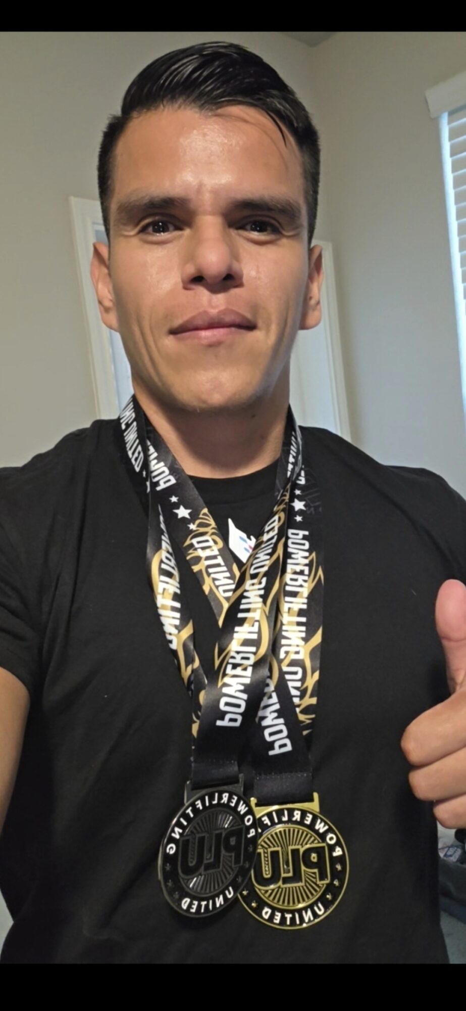 Person wearing medals around neck, giving thumbs up, indoors with window blinds in background.