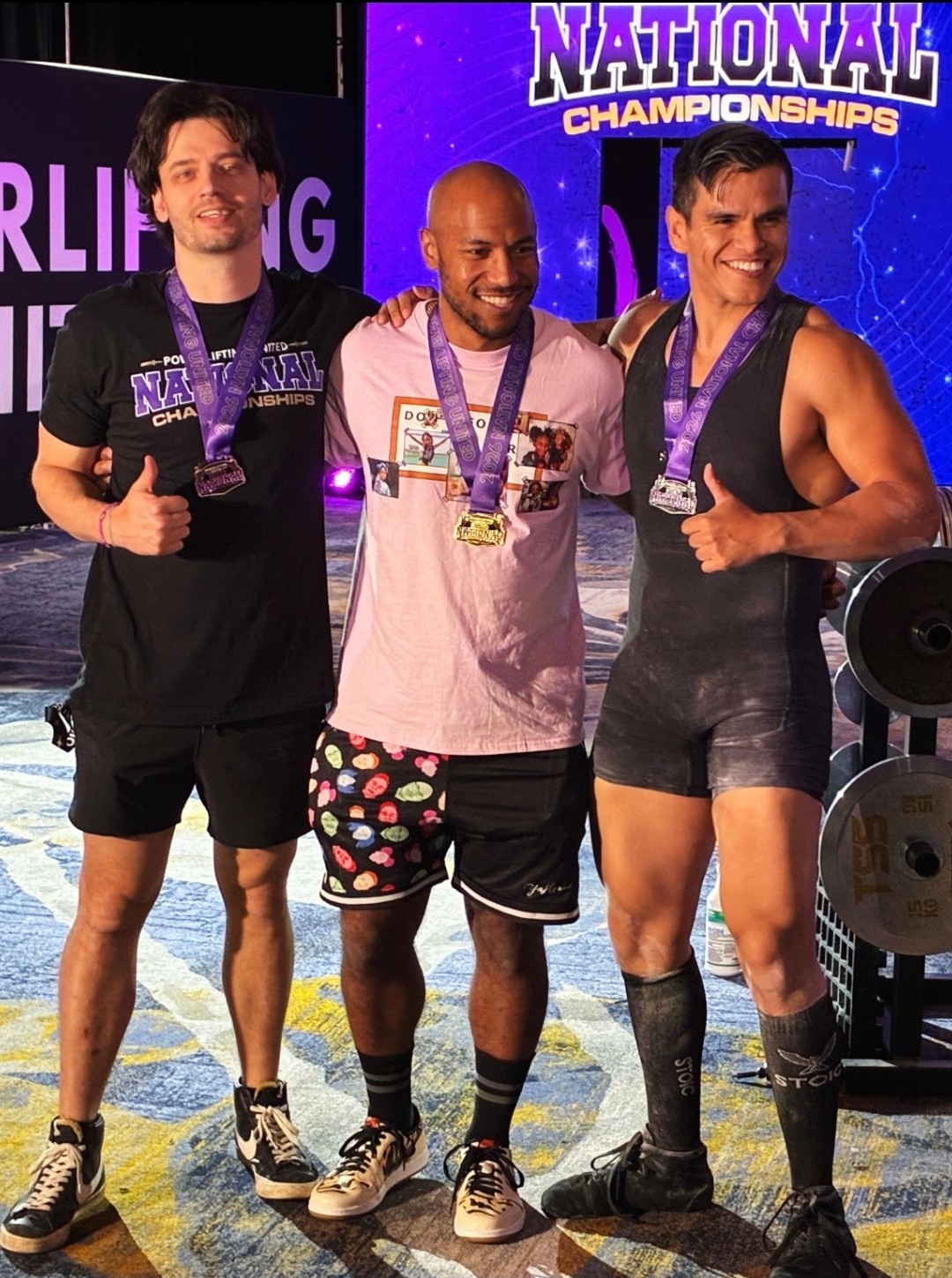 Three men with medals around their necks stand together, smiling and giving thumbs up at a championship event.