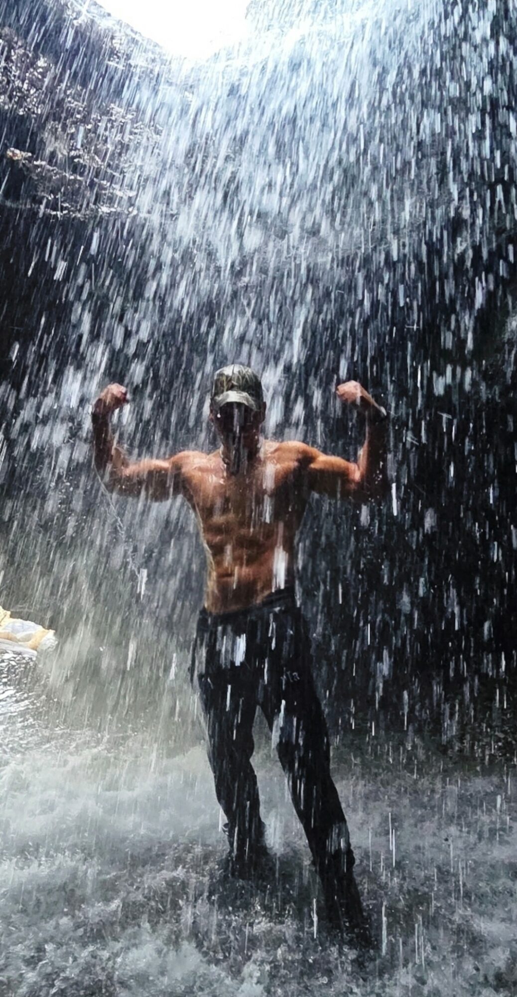 Person standing under waterfall with arms raised, water cascading around them, dark background, wet clothing, muscular build