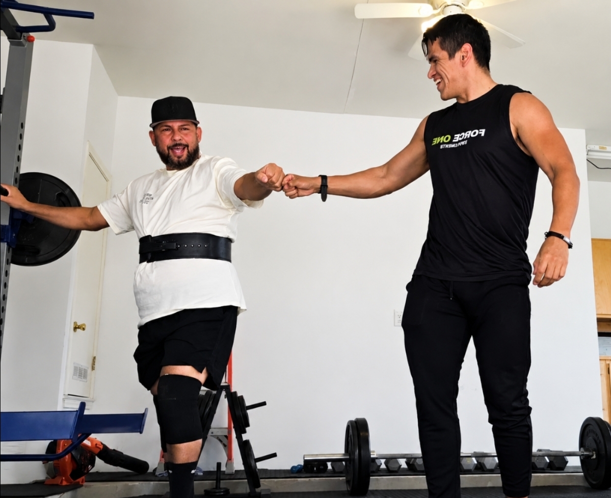 Two men shake hands in a gym, one wearing a weightlifting belt and knee brace, the other in workout clothes, smiling.