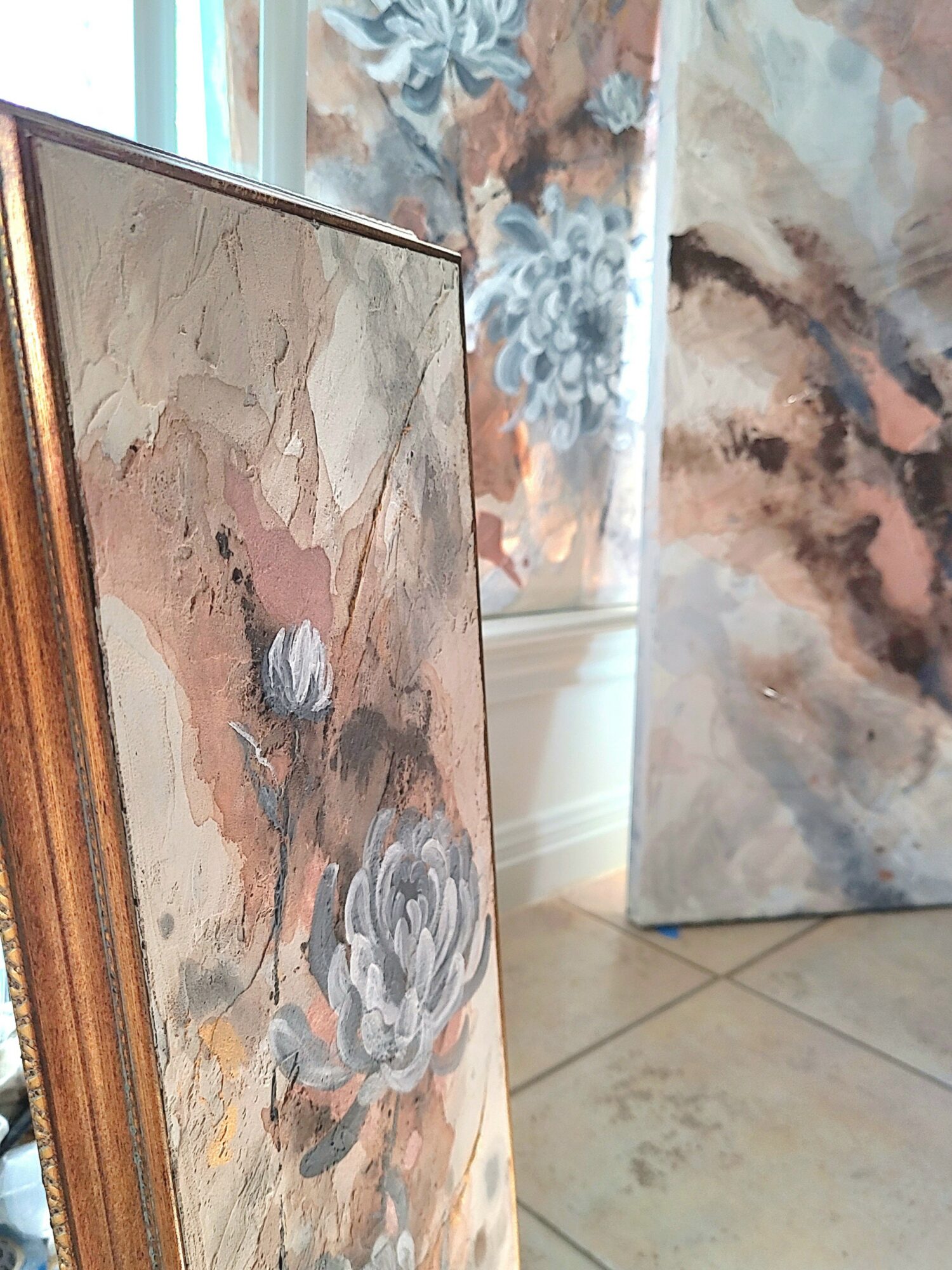 Framed abstract painting with floral elements leaning against a wall, with another artwork partially visible behind it.