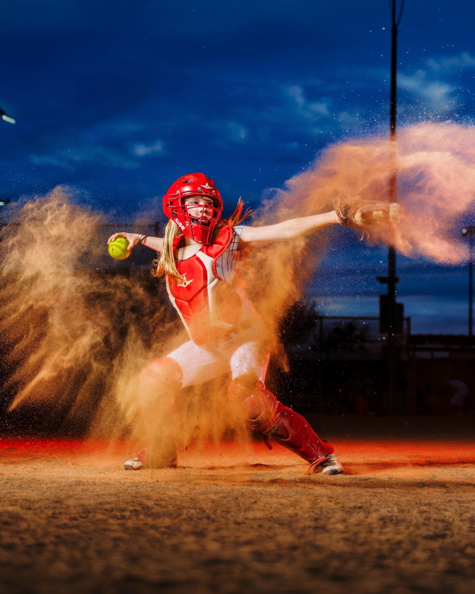 Baseball player in red uniform and helmet throws orange powder on the field during night game.