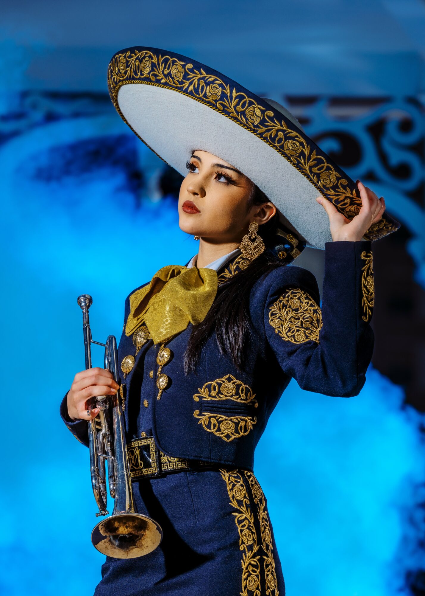 Woman in ornate costume with large hat, holding a trumpet, against a blue background.