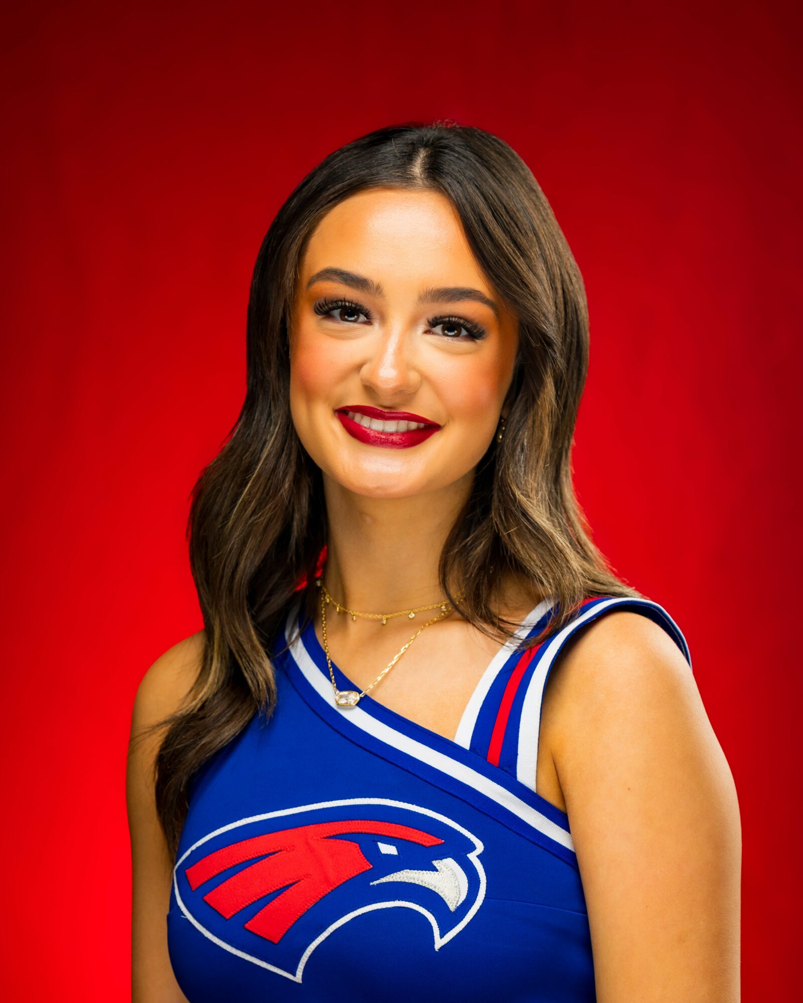 Young woman with long wavy hair, smiling, wearing a blue sports jersey with red and white accents, against a red background.