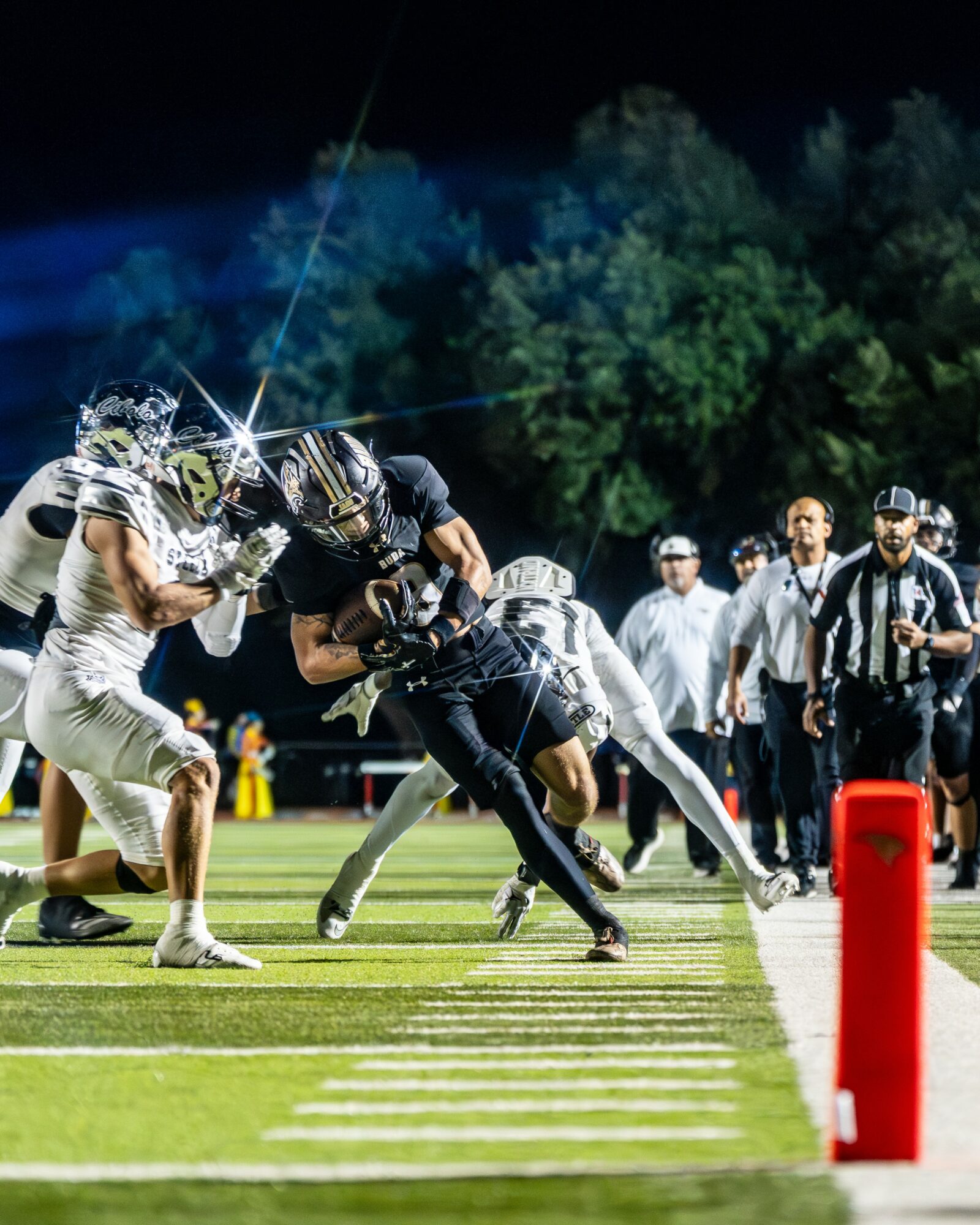 Football players in action during a game at night, with referees and spectators in the background on a lit field.