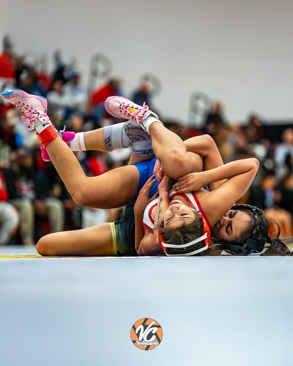 Two young girls wrestling on a mat with a crowd watching in the background, one girl pinning the other.