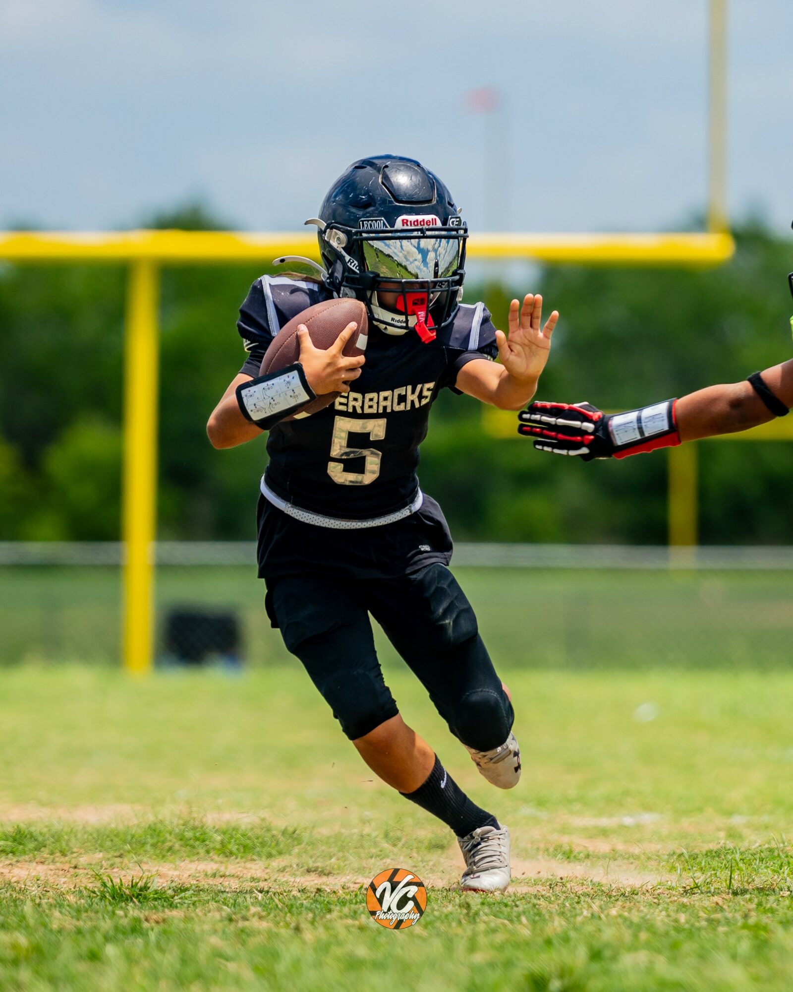 Young football player running on field with helmet and uniform, holding ball, reaching out to a teammate's hand, green field background.