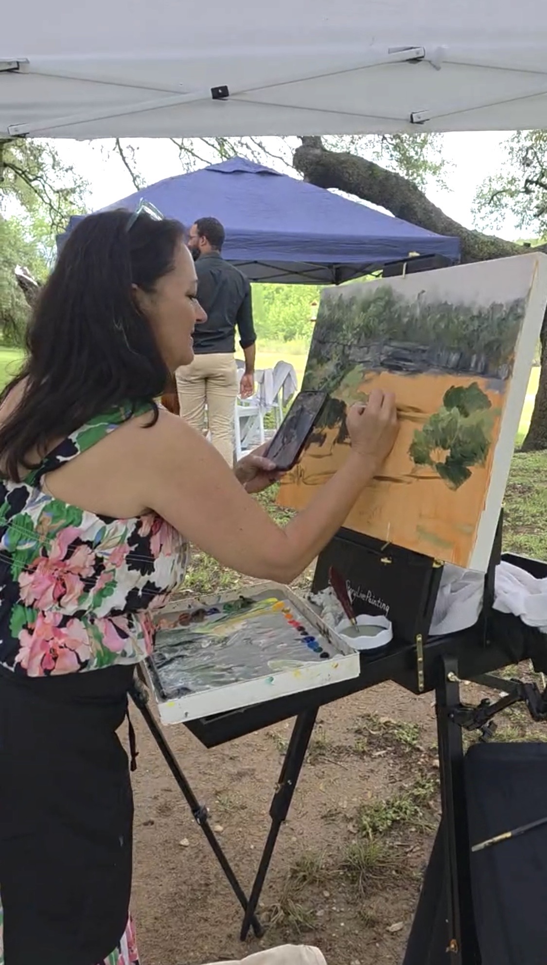 Woman painting outdoors under a canopy, with trees and people in the background.