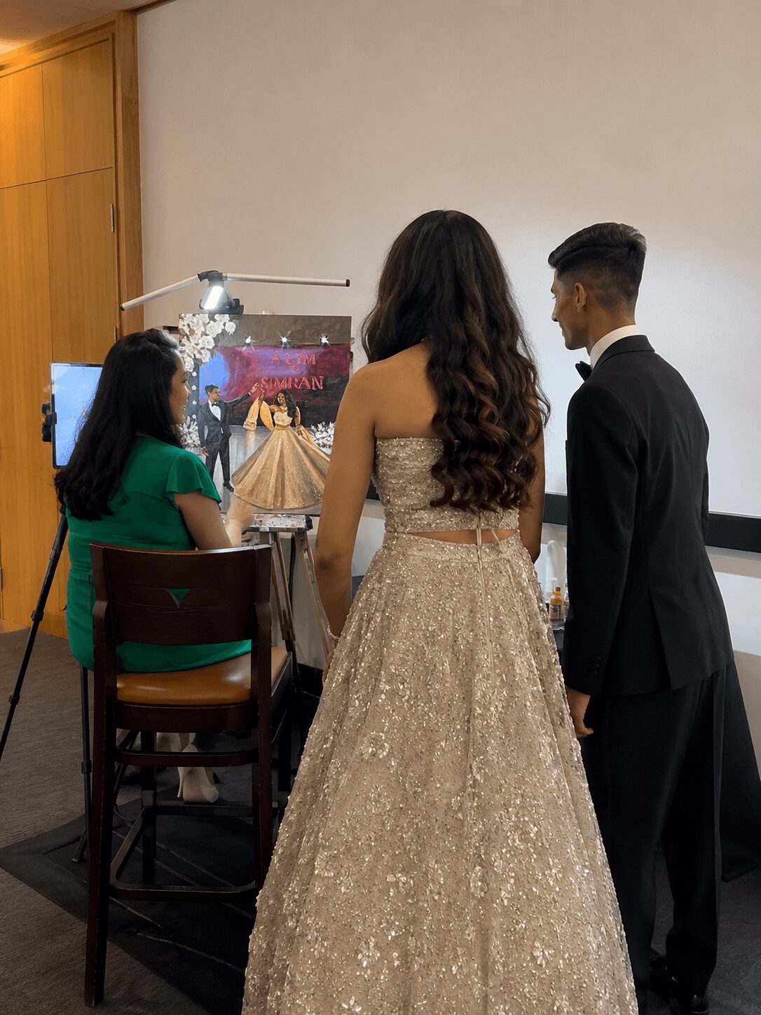 Three people in formal attire stand in a room with a plain wall, facing a screen displaying a wedding scene.