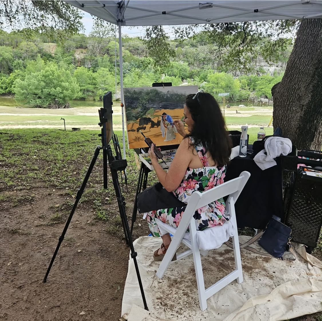 Woman painting outdoors under a canopy, seated on a white chair, with trees and a river in the background.