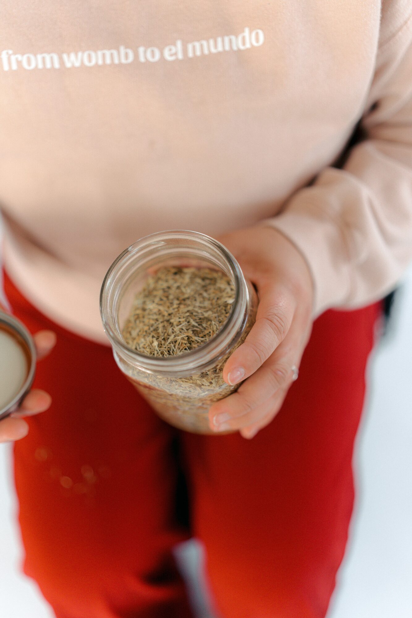 Person holding a small glass jar filled with dried herbs or spices, wearing a beige top and red pants.