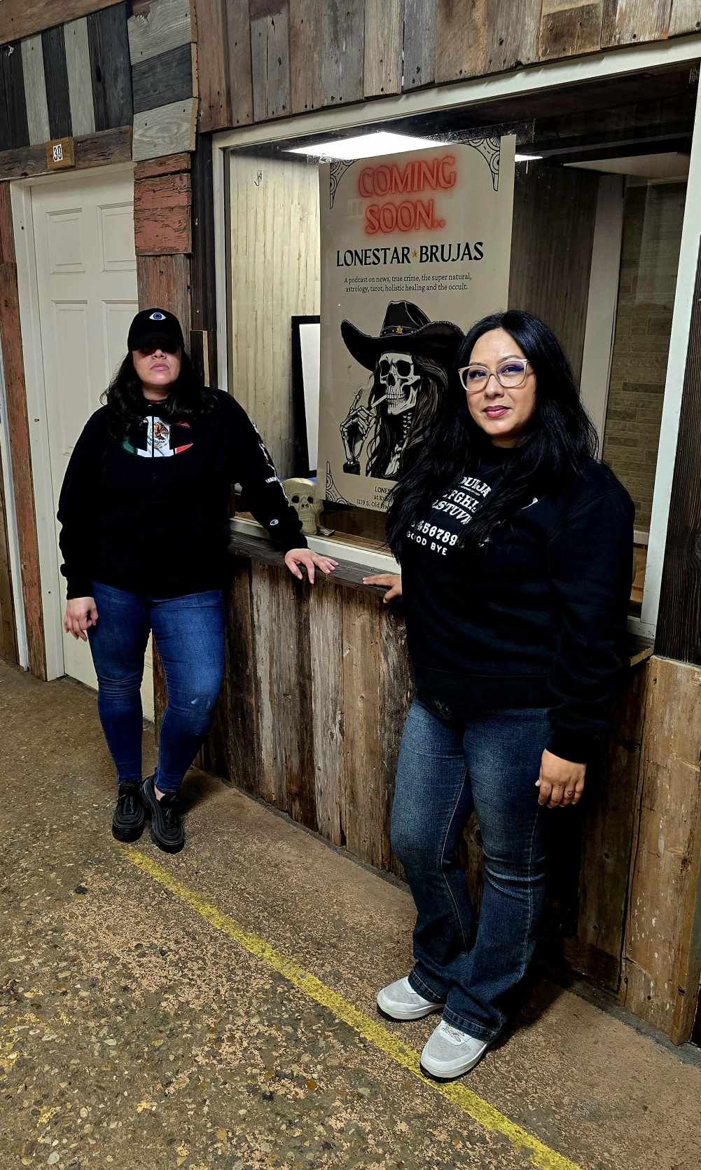 Two women stand inside a rustic wooden building, one touching a framed picture of a dog, both smiling.