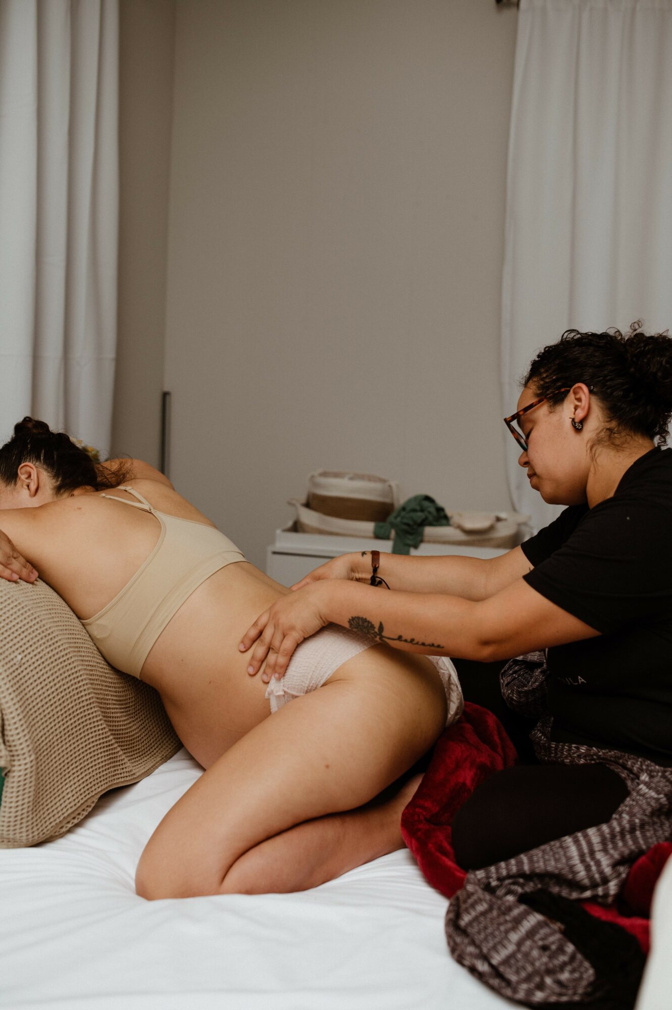 Person lying on bed with another person attending to them, in a room with white curtains and a stack of books.