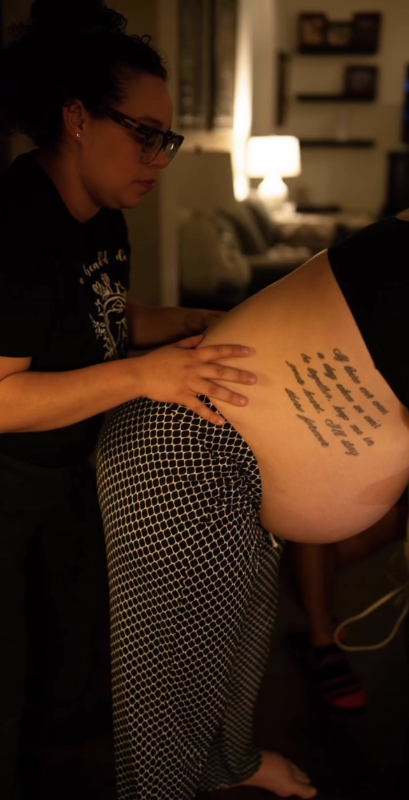 Person with glasses and dark hair leaning over a person's back with writing, in a dimly lit room.