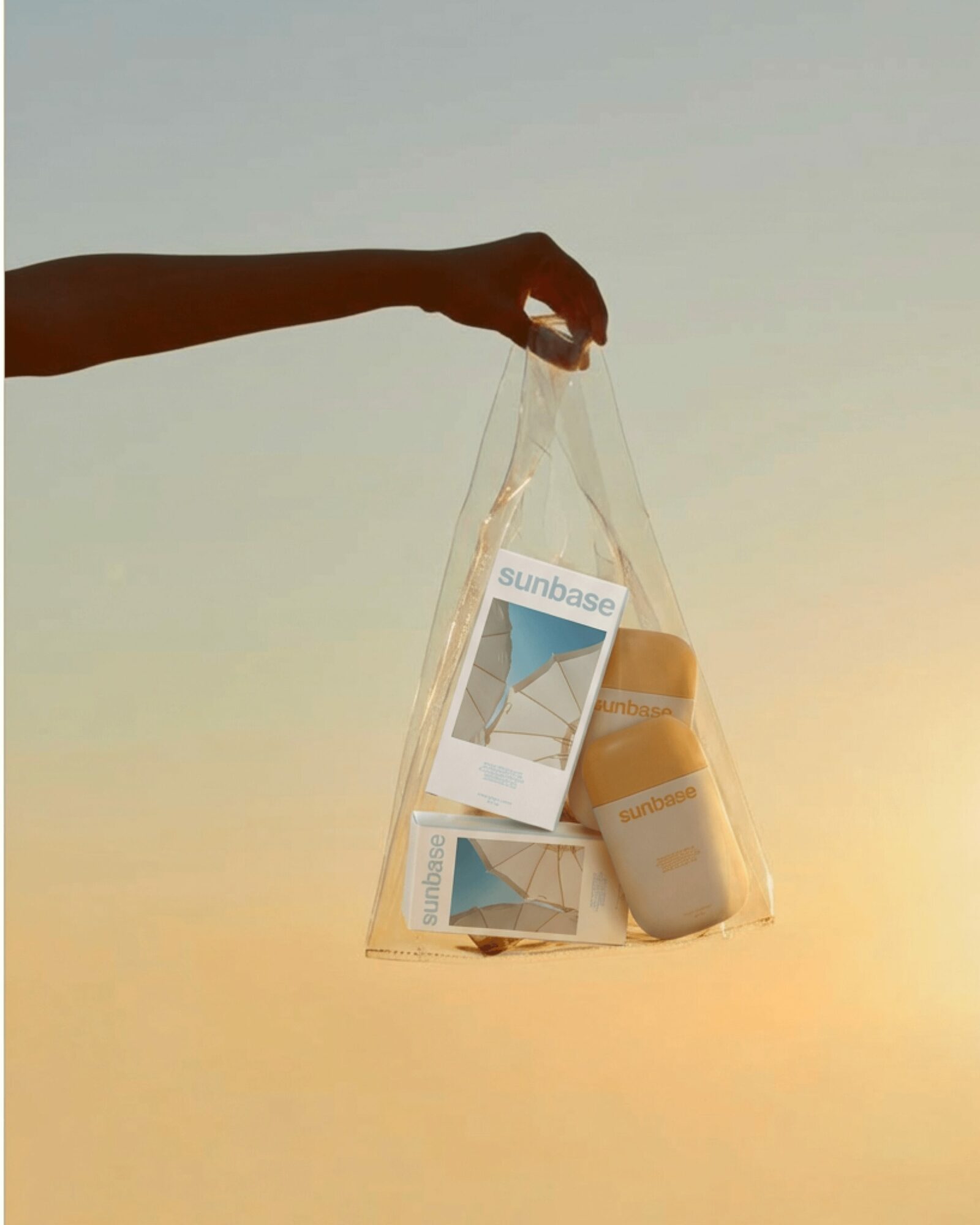 A hand holds a transparent bag containing sunscreen and a brochure against a sunset sky, with a gradient of warm colors.