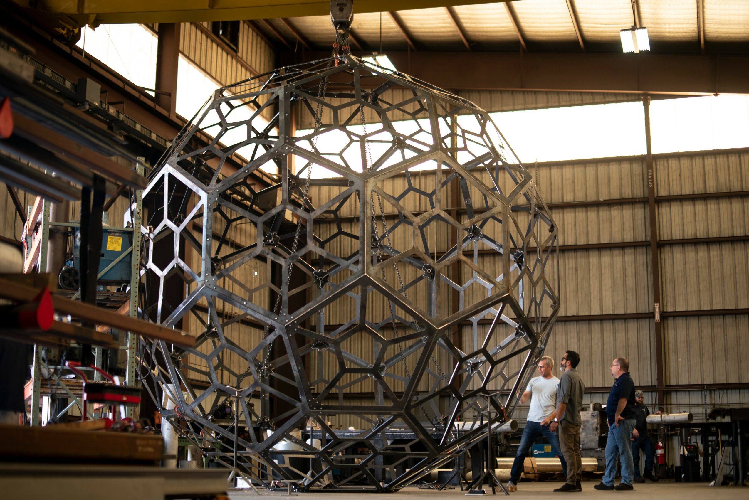 Large geometric metal sphere structure inside a warehouse with people walking nearby.