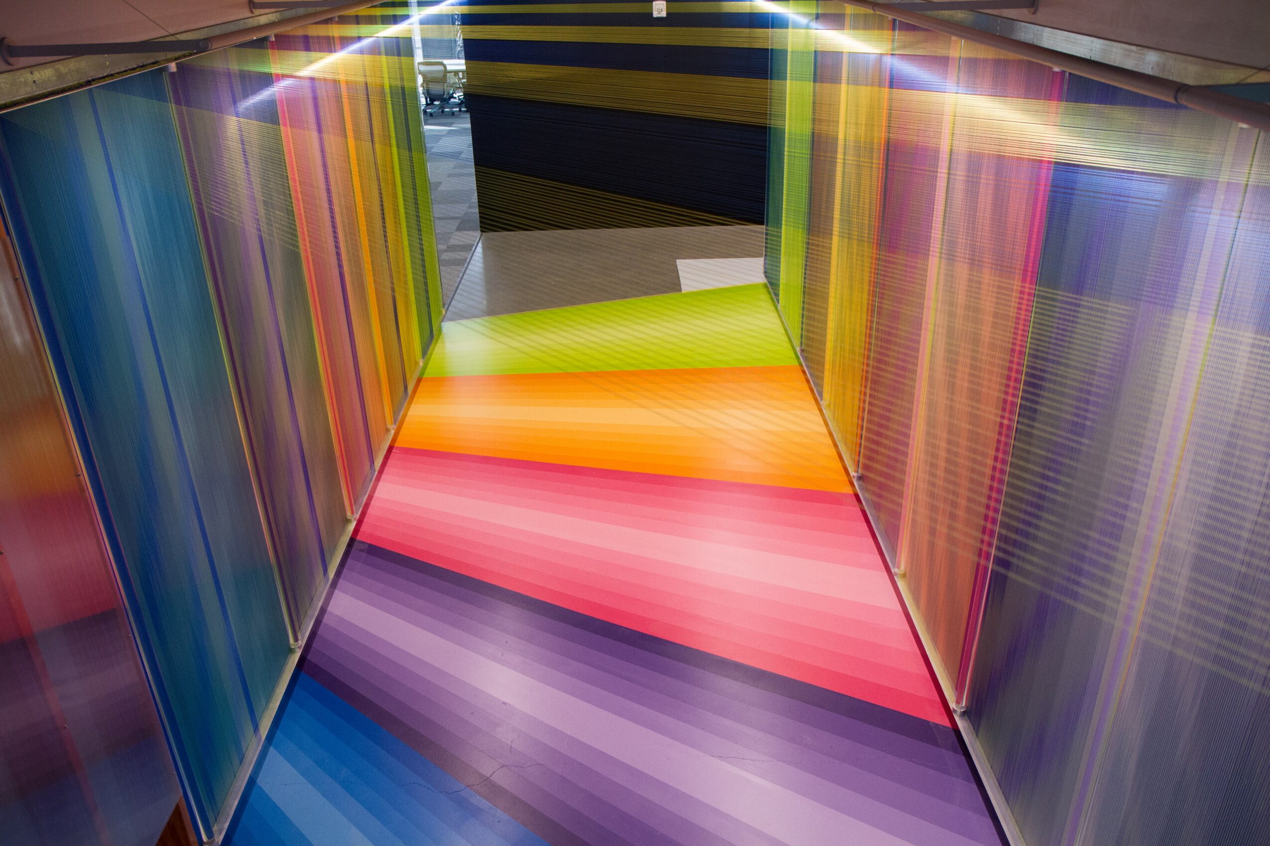 Colorful corridor with rainbow-colored floor and reflective walls, leading to a bright area with a green seat, viewed from the side.