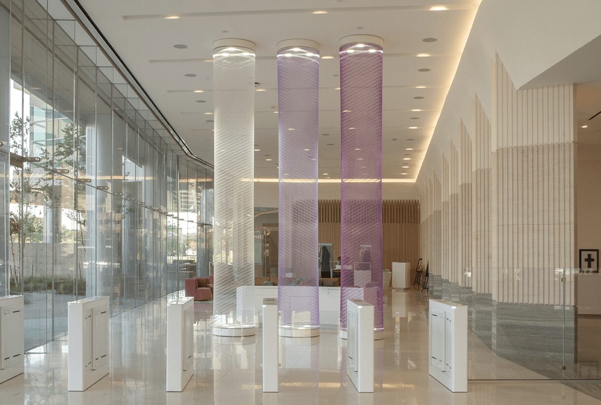 Interior space with three tall, illuminated columns in white, purple, and pink, and glass walls with reflections.