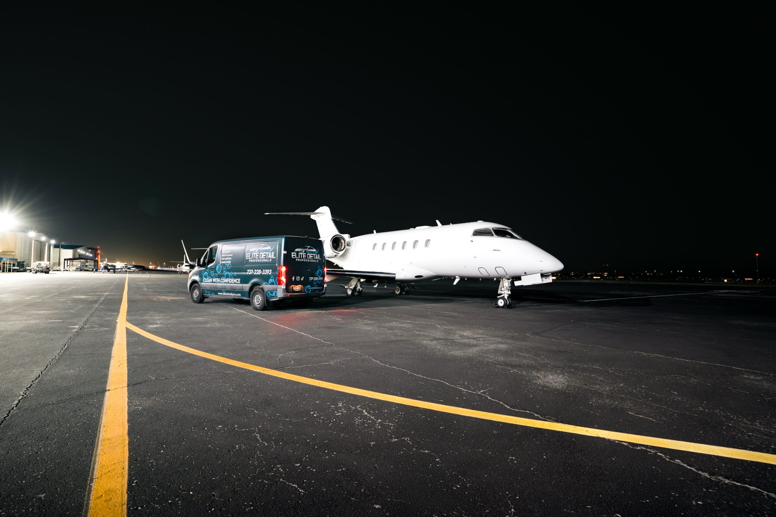 Night scene with a white private jet and a blue van parked on the tarmac, illuminated by airport lights.
