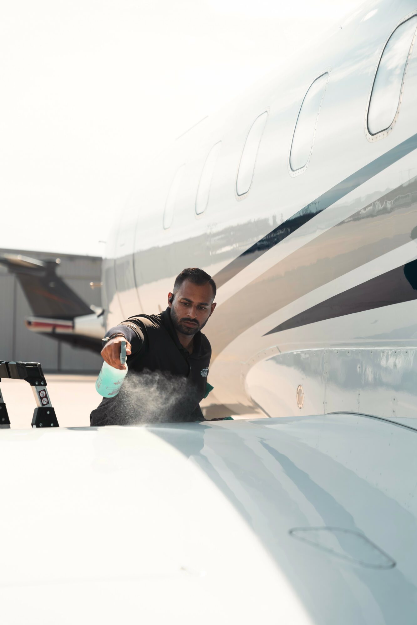 Man cleaning the wing of a private jet with a cloth, airplane parked outdoors, clear sky in background.